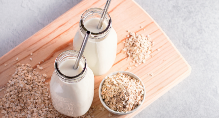 Two bottles of milk with straws on a wooden board, surrounded by oats and a bowl of granola.